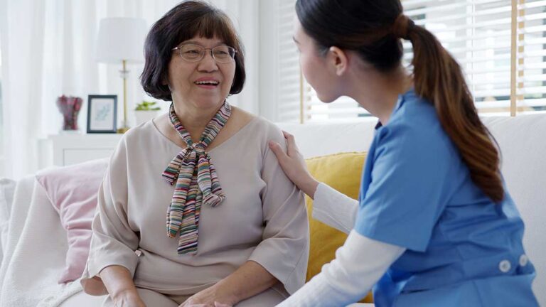 Family members visit an older adult in a bright assisted living common room while a staff member checks visitors in.